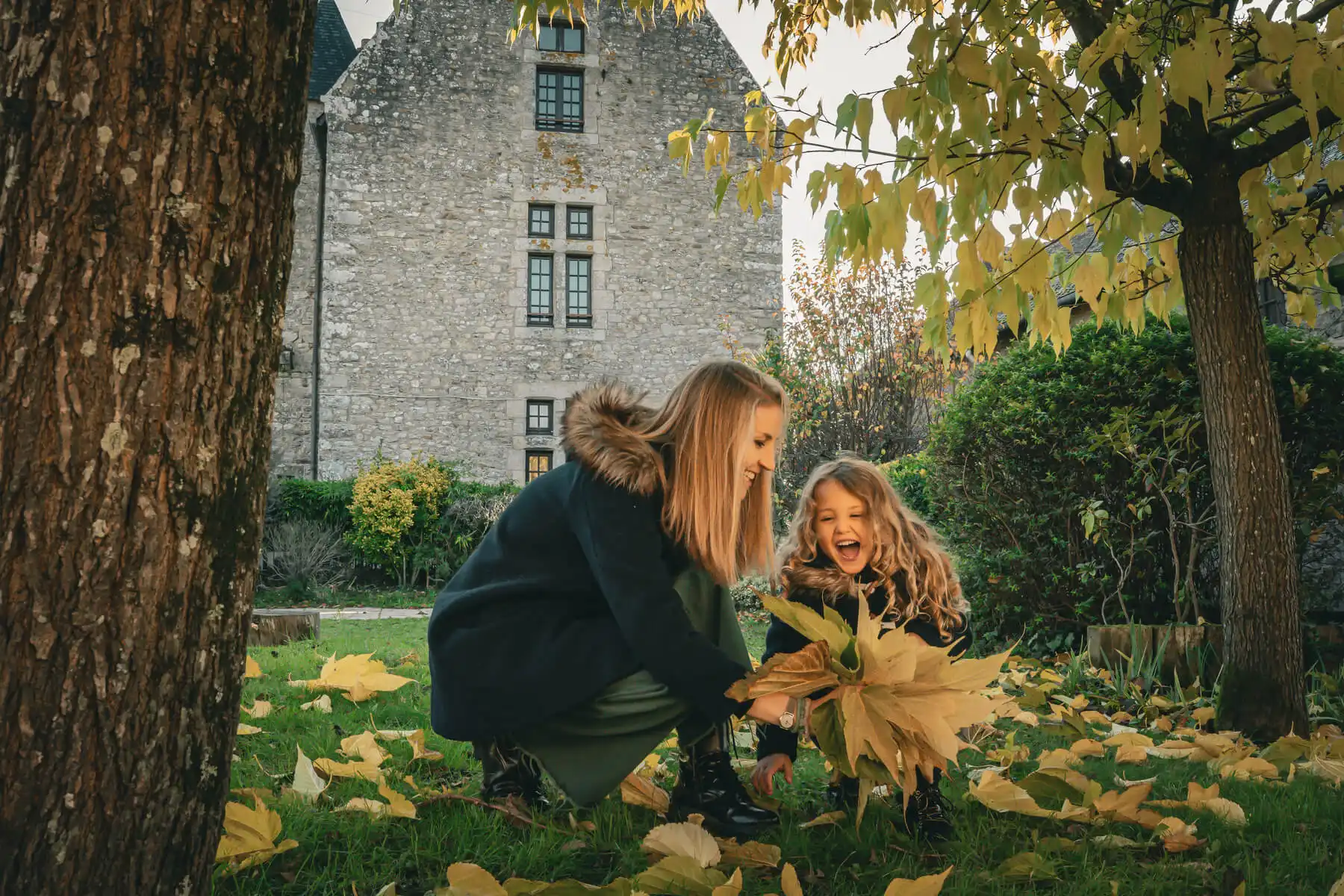 Mère et enfant jouant avec des feuilles sous un arbre dans le jardin de la Maison OBONO