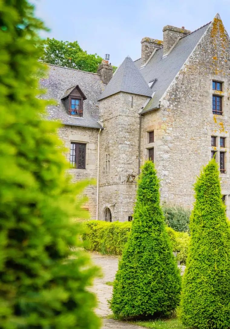 Façade en pierre du Manoir de Kerdréan vue depuis le jardin, entourée de cyprès et végétation verdoyante, Maison OBONO, Le Bono, Bretagne