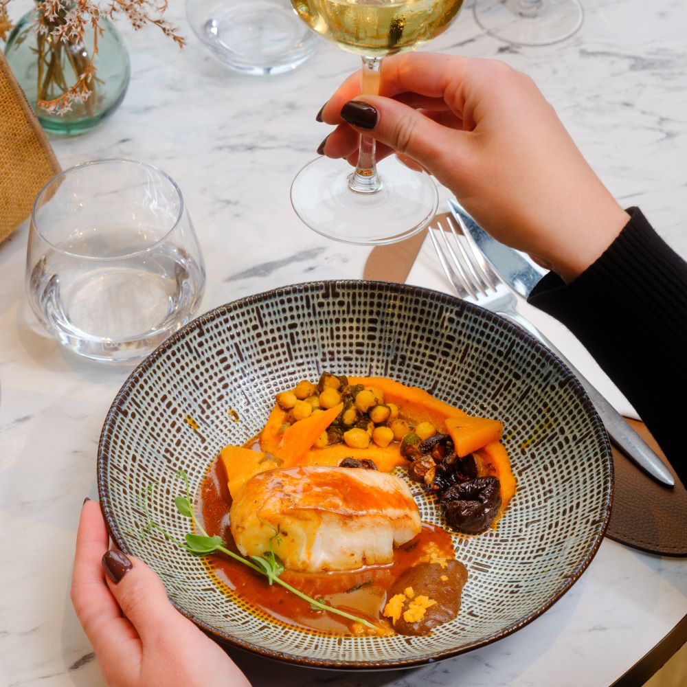 Plat de poisson blanc sur lit de légumes mijotés, servi dans un bol artisanal à motifs, accompagné d'un verre de vin blanc
