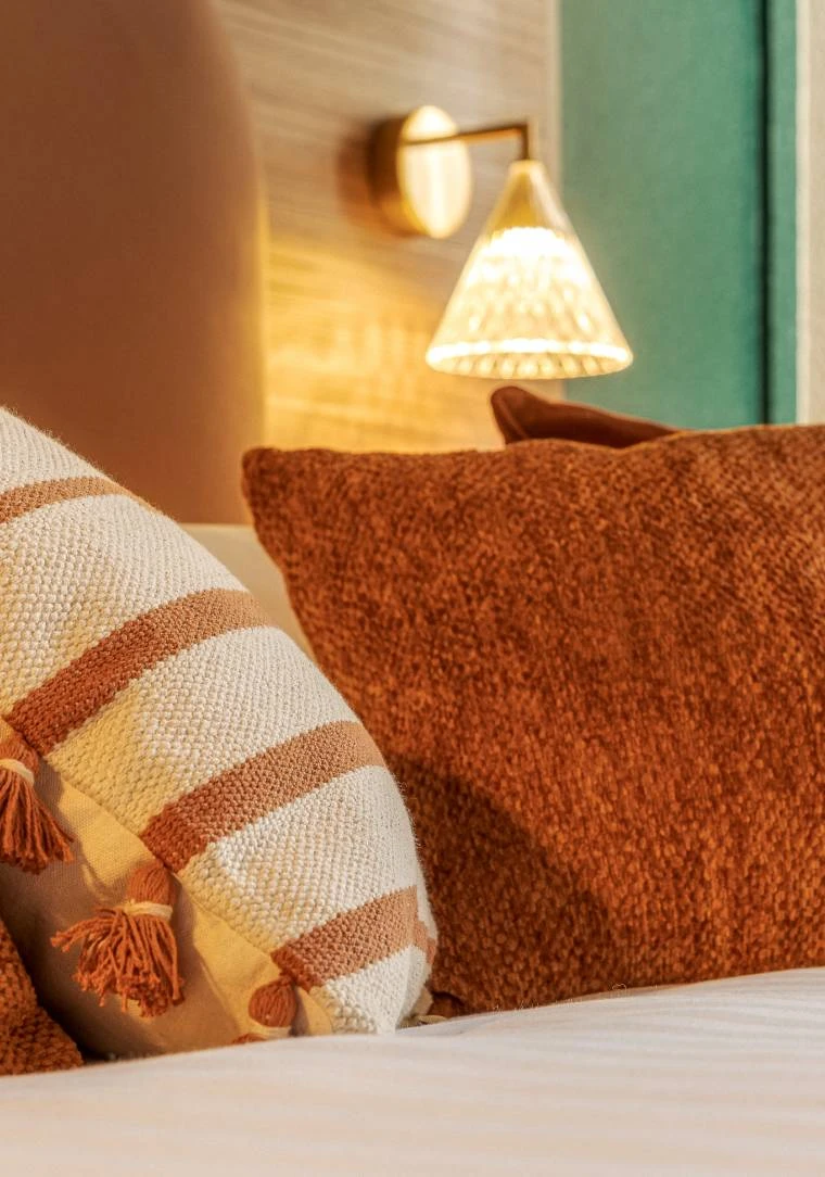 Coussins décoratifs terracotta et rayés sur lit avec applique murale dorée dans une chambre de la Maison OBONO