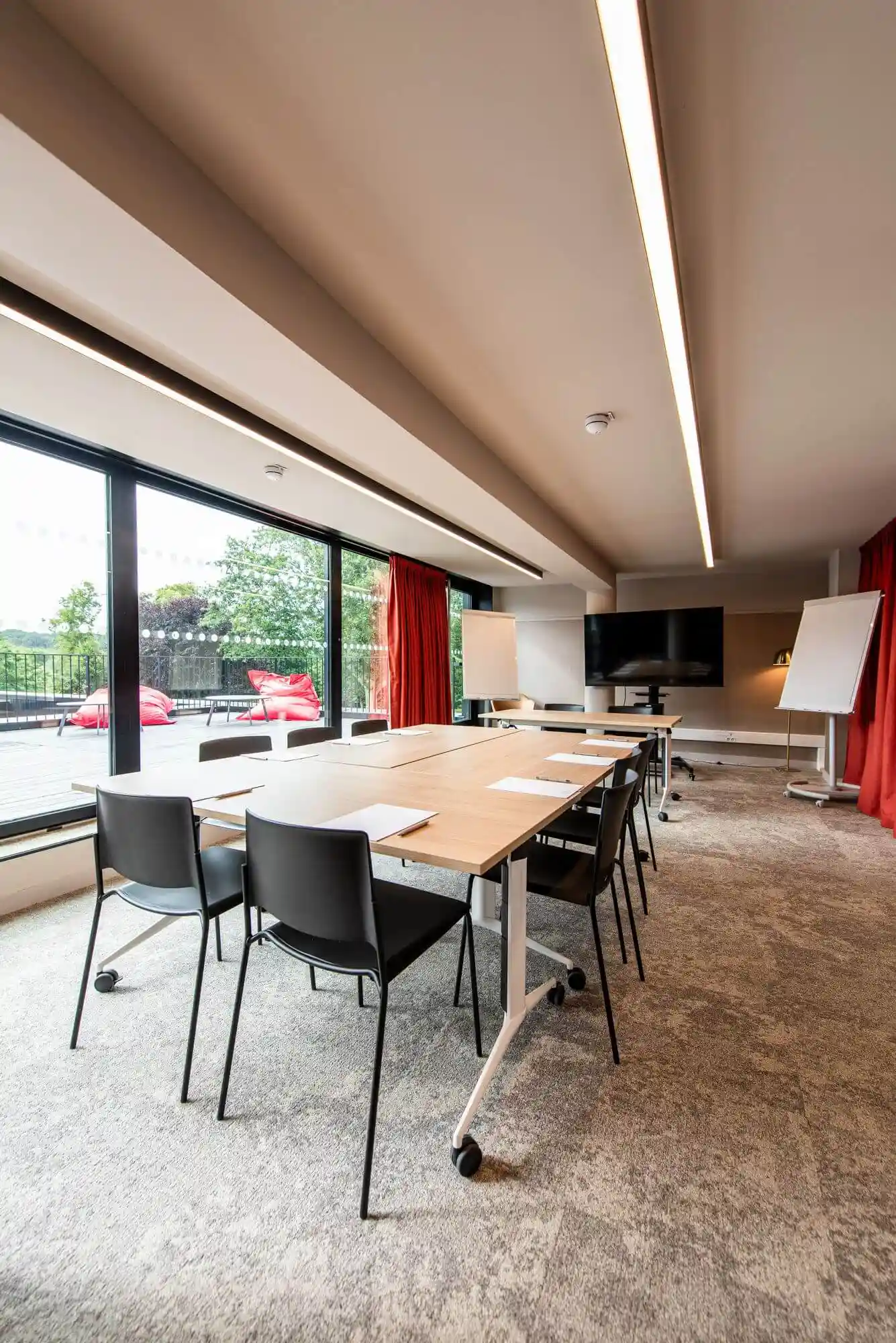 Salle de séminaire modulable de la Maison OBONO : tables en configuration réunion, chaises noires, écran TV, paperboard, baies vitrées sur terrasse et rideaux rouges